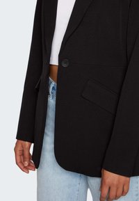 Černý blazer s texturovanou tkaninou, zapínání na jeden knoflík a klopovým límcem. Obsahuje přední kapsu a volný střih, kombinovaný se světle modrými džíny.