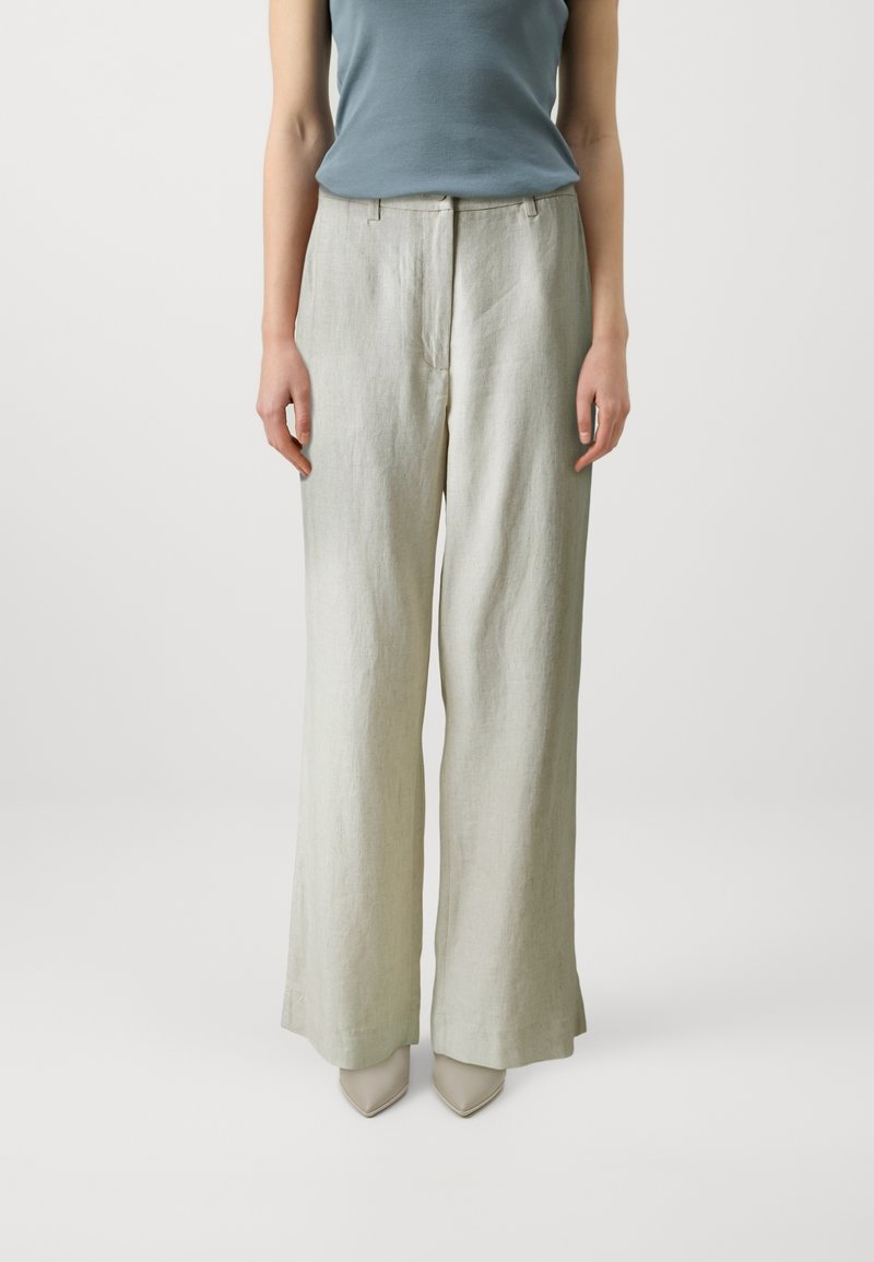 Pantalons larges en lin vert clair avec une texture lisse, dotés d'une fermeture éclair à l'avant et de détails de plis. Associés à des escarpins beiges à bout pointu.