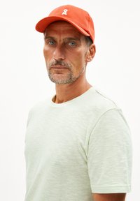 Helles grünes, strukturiertes T-Shirt mit rundem Ausschnitt, kombiniert mit einer orangefarbenen Mütze mit einem weißen Logo. Der Hintergrund ist schlicht weiß.