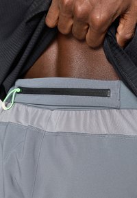 Γκρι αθλητικές βερμούδες με υφασμάτινη ζώνη, που διαθέτουν τσέπη με φερμουάρ μαύρου χρώματος και πράσινο λαιμό. Λείο ύφασμα.