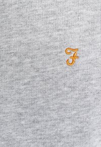 Tela gris con una textura suave y un patrón jaspeado sutil. Cuenta con una letra "F" naranja bordada como detalle destacado.