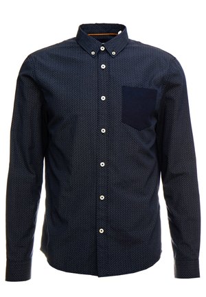 Chemise à manches longues bleu marine avec un petit motif à pois blancs, col boutonné, et une poche plaquée sombre sur le côté gauche de la poitrine. Boutons blancs.