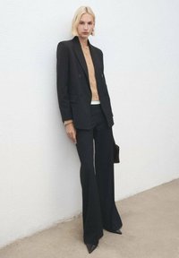 Blazer negro a rayas finas sobre una blusa beige con botones, combinado con pantalones negros de campana y tacones afilados negros, sosteniendo un bolso pequeño.
