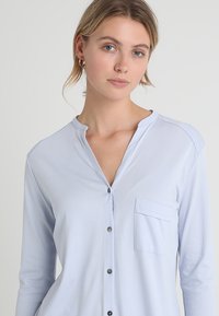 Světle modrá košile s dlouhým rukávem, knoflíkovým zapínáním, kulatým límečkem a jednou náprsní kapsou. Měkká tkanina s hladkým povrchem a jemným leskem.