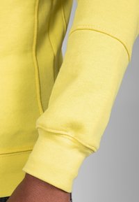 Sudadera amarilla con puños acanalados, que presenta una tela texturizada y detalles de costura visibles a lo largo de las costuras, mostrando un primer plano del brazo.