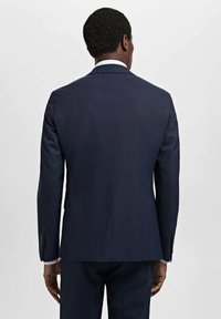 Mörkblå kavaj i ull, skräddarsydd design, två bakventiler, enstaka knappstängning, textil med struktur och silverknappdetaljer.