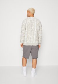 Light gray textured sweatshirt with intricate patterns, paired with dark gray shorts and white sneakers. Model facing away from the camera.