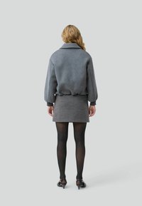 Una donna con capelli ondulati sta in piedi di spalle, indossando una giacca grigia, una mini gonna a fantasia e collant neri con scarpe con tacco.