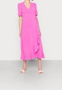 Vestido envolvente rosa con escote en V, mangas cortas abullonadas, dobladillo con volantes y detalle de lazo en el costado; combinado con sandalias de tacón beige.