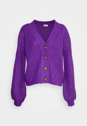 Violetti paksu neulejakku, jossa pitkät pallohihat ja etupuolella neljä ruskeaa nappia, V-kauluksella.