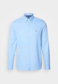 Chemise bleu clair à manches longues avec des boutons, un col boutonné et un petit logo brodé sur la poitrine gauche, présentée sur fond blanc.