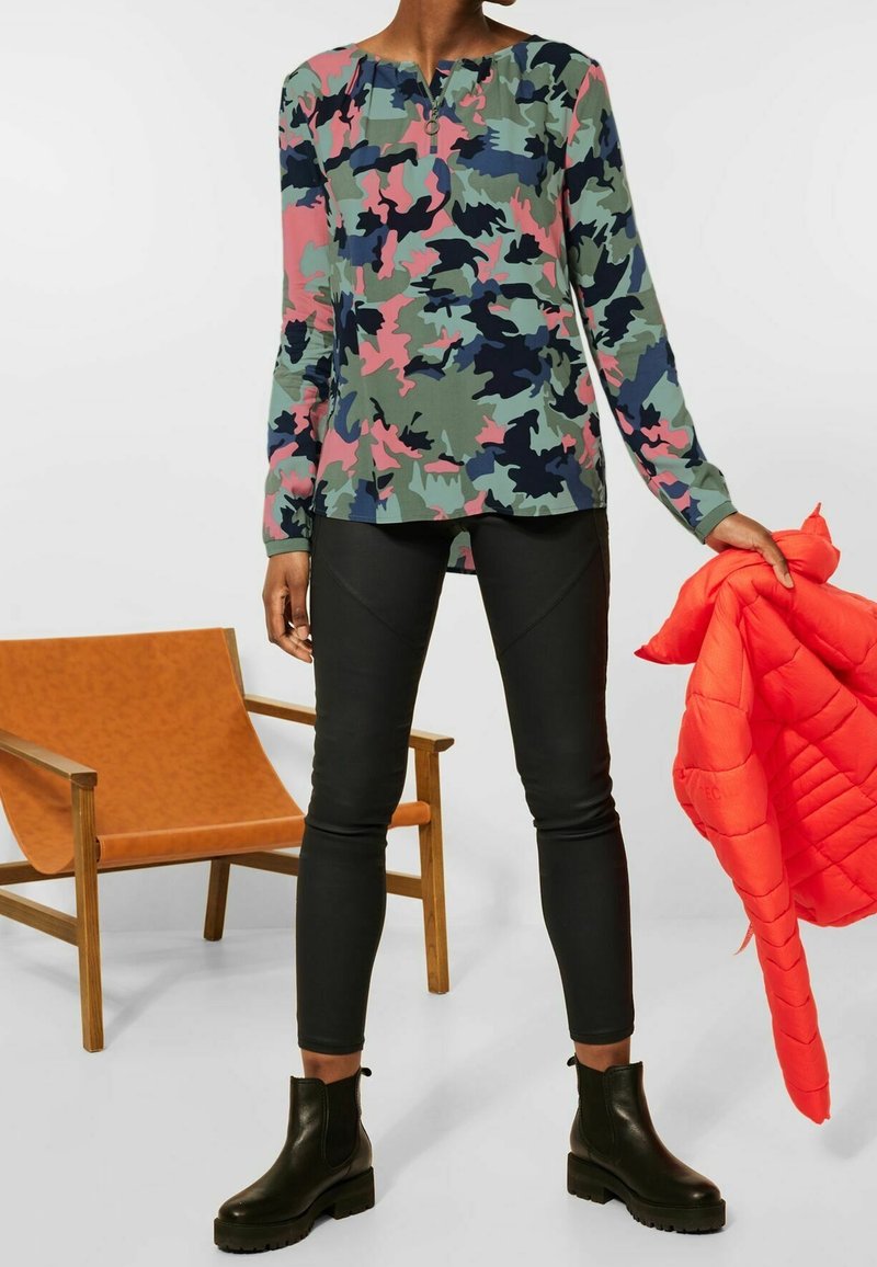 Personne portant un haut à manches longues imprimé camouflage, un pantalon noir, des bottes noires, et tenant une doudoune orange vif à côté d'elle.