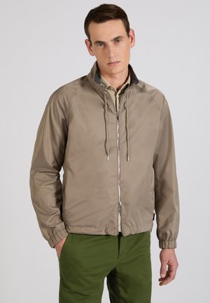 Junger Mann trägt eine beige Jacke mit Reißverschluss und Kordelzug sowie grüne Hose, steht mit einer Hand in der Tasche vor schlichter Hintergrund.