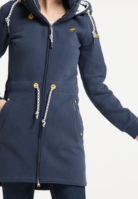 Marineblå fleeceskjorta med dragkedja fram, justerbar huva, vita dragsko och gula detaljer; har två sidofickor och logodetalj.