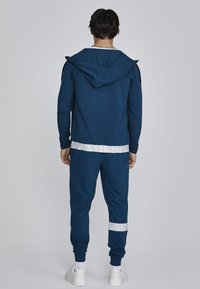 Marineblauer Trainingsanzug mit Kapuze, ausgestattet mit einem weißen Band an der Taille und den Bündchen, glatter Textur und gerippten Akzenten an Ärmeln und Beinen.