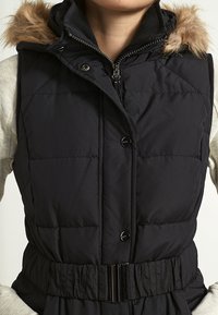 Kobieta ubrana w czarną pikowaną kamizelkę bez rękawów z kapturem wykończonym futrem, metalowymi napami, zamkiem błyskawicznym oraz szerokim elastycznym paskiem w talii.