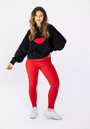 Czarny sweatshirt z graficznym motywem czerwonej szminki, zestawiony z dopasowanymi czerwonymi legginsami. Modelka nosi białe sneakersy, prezentując casualowy strój.