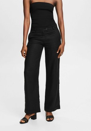 Vrouw draagt een zwart strapless top, wijde zwarte broek en zwarte open-toe sandalen met blokhak, staand tegen een effen achtergrond.