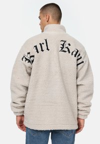 Beige sherpa fliisjakker kõrge kaelusega. Seljal must broderitud tekst "Brl Kay". Looduslik lõige, tekstureeritud pind ja pikad varrukad.