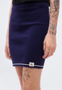 Πλεκτή μίνι φούστα σε σκούρο μοβ με υφασμένο σχέδιο καλωδίου. Διαθέτει ριμπ ζωνάρι στη μέση και λευκές ρίγες στο τελείωμα. Φέρει ορατό λογότυπο.