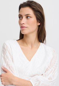 Weißes Kleid mit V-Ausschnitt und transparenten, strukturierten Ärmeln, die ein Wellenmuster aufweisen. Glatter Stoff mit sanften Akzenten, figurnah an der Taille.