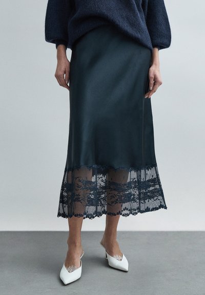 Jupe en satin bleu sarcelle foncé avec une bordure en dentelle transparente ornée de motifs floraux à l'ourlet. Associée à des chaussures blanches à talons pointus.