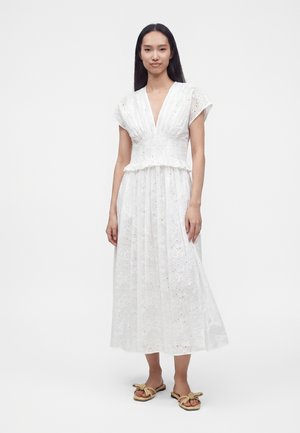 Femme portant une robe blanche à manches courtes, longueur cheville, avec des motifs floraux découpés et des sandales beiges, debout devant un fond uni.