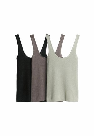 Frau trägt ein beige geripptes Tanktop, lockere hellblaue Jeans, schwarze Riemchensandalen mit Absatz, steht mit einer Hand in der Hüfte vor weißem Hintergrund.