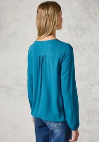 Blusa a maniche lunghe color teal con un motivo testurizzato, scollo rotondo e dettaglio raccolto sul retro. Abbinata a pantaloni di denim.