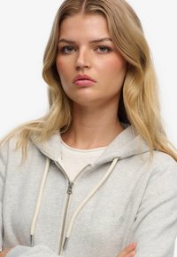 Grijze hoodie met rits van zachte stof, met een gestructureerd ontwerp, witte trekkoorden en een subtiel logo geborduurd op de borst.