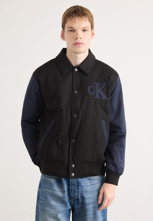 Zwarte en marineblauwe varsityjack met een knopenfront, zijzakken, ribgebreide boorden en zoom, voorzien van een "CK" logo op de borst.