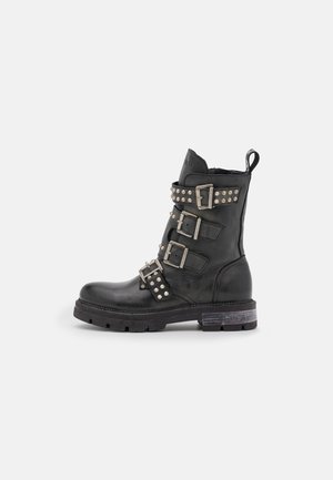 Black leather combat boot with four studded buckle straps and thick tread sole, viewed from the side on a plain background.