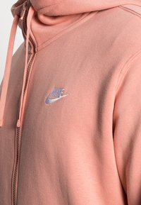 Närbild av en rosa zip-up hoodie med en vit broderad Nike-logotyp på bröstet och matchande rosa dragskor på huvan.