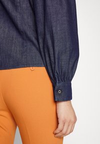 Camicia di denim blu marina con polsini a maniche arricciate, con bottone decorativo. Abbinata a pantaloni arancioni aderenti, che mettono in mostra una texture di tessuto liscia.