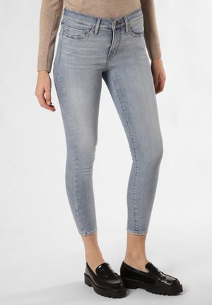 Femme portant un jean skinny bleu clair, un haut beige à manches longues et des mocassins noirs épais, debout sur un fond blanc.