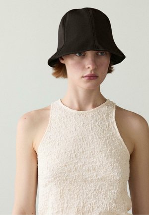 Ung person med kort hår iført en tekstureret hvid ærmeløs top og en sort bucket hat, der kigger let mod højre for kameraet.