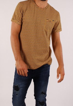 Mann in senfgelber gemusterter Kurzarm-T-Shirt und dunkelblauen zerrissenen Jeans, der vor einem einfarbigen Hintergrund steht.