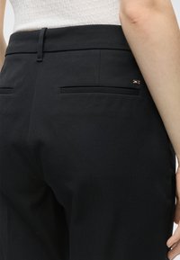 Κοντινό πλάνο μαύρου προσαρμοσμένου παντελονιού με πίσω τσέπες welt και ένα μικρό ορθογώνιο λογότυπο στην περιοχή της δεξιάς τσέπης, φορεμένο με λευκό υφασμάτινο τοπ.