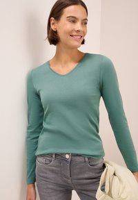 Langärmliges, V-Ausschnitt-Shirt aus tealfarbener Baumwolle, kombiniert mit grauen Hosen. Der Stoff wirkt glatt und hat eine lässige Passform. Eine leichte Jacke ist über den Arm gelegt.