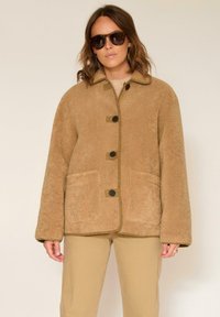 Veste polaire beige avec col, cinq boutons et deux poches avant. Bordures contrastantes autour des bords, avec une finition douce et texturée.