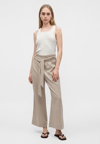 Mujer joven con camiseta sin mangas blanca con borde festoneado, pantalones anchos de talle alto color beige con cinturón de lazo, y sandalias negras tipo chanclas, de pie.