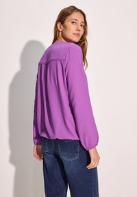 Lilla langærmet bluse med tekstureret stof, elastiske manchetter og en plisseret rygdesign. Kombineret med mørkeblå jeans med en klassisk skæring.