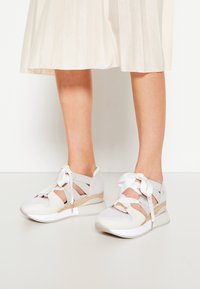 Baskets blanches avec un talon compensé tissé, présentant un mélange de matériaux texturés, un laçage à l'avant avec des accents en forme de nœud et des détails découpés sur les côtés.