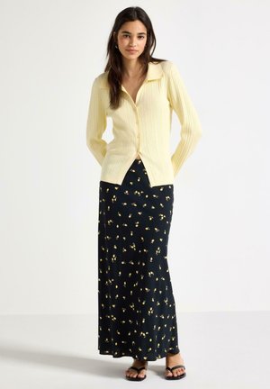 Ung kvinde iført en lysegul ribbet cardigan med knapper og et langt sort skørt med et lille gult blomsterprint, stående med hænderne bag ryggen.
