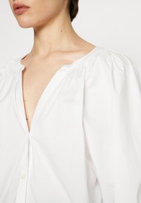 Weiße Baumwollbluse mit tiefem V-Ausschnitt, gerafftem Schulterdetail und Knopfverschluss. Glatte Textur mit legerer Passform.