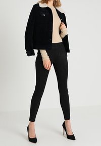 Giacca nera in suede con colletto in pelliccia bianca, maglietta attillata beige a maniche lunghe, pantaloni skinny neri e tacchi alti neri.