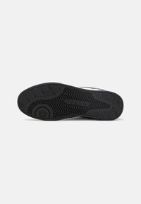 Schwarze Sneaker-Sohle mit strukturiertem Griffmuster, das ein kreisförmiges Logo und eine geprägte Markenkennzeichnung aufweist. Das Material wirkt gummiert für Langlebigkeit.