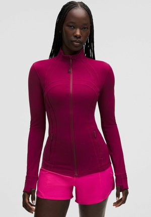 Mujer que lleva una chaqueta magenta ajustada con cremallera y manga larga y pantalones cortos deportivos de color rosa brillante, de pie frente a un fondo liso.