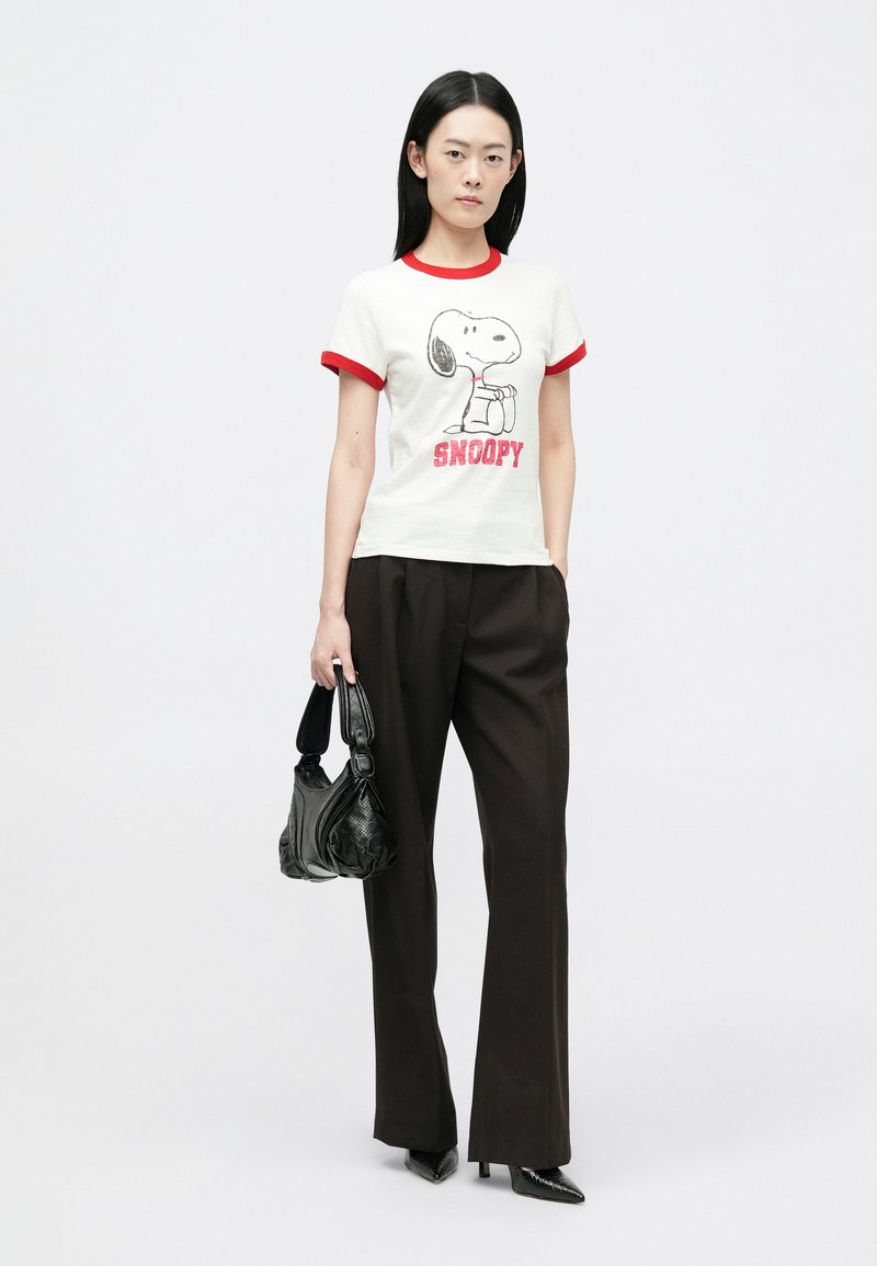 T-shirt blanche avec des bordures rouges présentant un graphique de Snoopy ; le mannequin porte un pantalon noir ample et tient un sac noir à finition texturée.