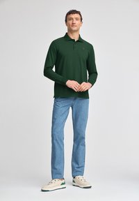 Grünes Langarm-Polohemd mit Kragen und kleinem gesticktem Logo, kombiniert mit hellblauen Jeans und beigen Sneakers.